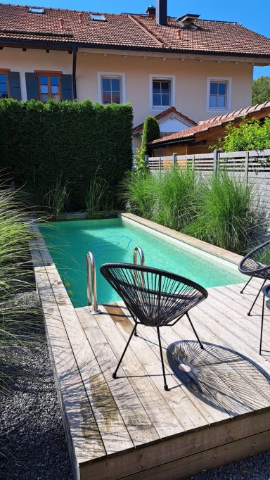 Holzterrasse mit einem Pool und zwei Stühlen, umgeben von grünen Pflanzen.